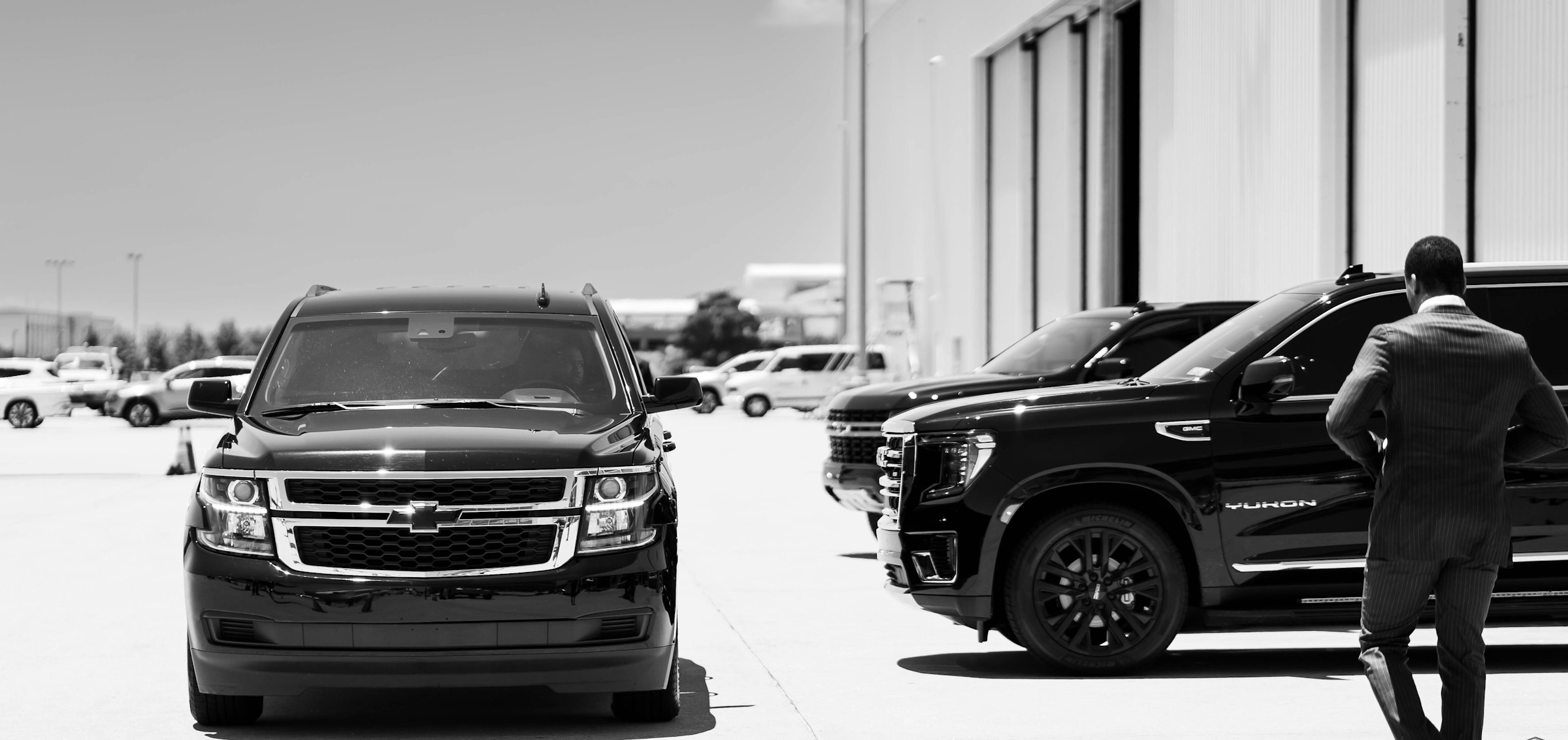 Chauffeur in a suit walking past a row of black SUVs, black and white
